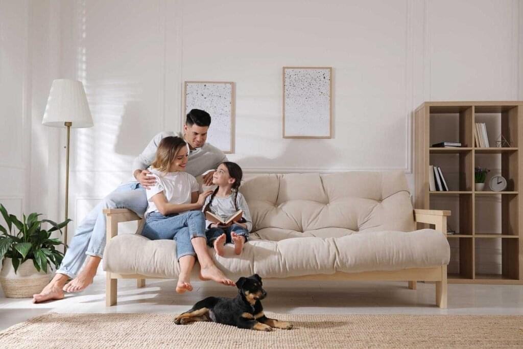 A mother and father relax on the couch reading with their young daughter, a puppy sits at their feet